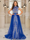 Woman wearing a Sparkling Royal Sapphire blue evening gown featuring a high neck, sleeveless bodice with intricate silver crystal beading, and a voluminous sheer chiffon overskirt in an elegant interior setting.